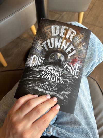 The media file shows a close-up, high-angle view of a person's lap, wearing light blue denim jeans. They are holding a book with a black cover featuring a tunnel-like image. The title of the book, written in large white letters, is "DER TUNNEL". Below this, the author's name, "CHRIS GEORGE", is also visible, along with the subtitle "NUR EINER KOMMT ZURÜCK" and the genre "THRILLER". Some of the text is partially obscured by the person's hand, which has neatly manicured nails. The background consists of a blurred view of a neutral-colored chair and wooden flooring. The lighting suggests it is daytime. The image appears to be taken indoors, possibly in a waiting area or a casual setting.