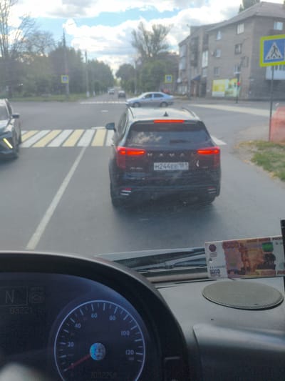 The image depicts a street view from inside a vehicle, likely a bus, in Alchevsk, Ukraine. The foreground shows a dashboard with a speedometer indicating approximately 20 km/h and a gear selection display showing "N". To the right of the dashboard, a 5000 ruble banknote is visible, propped up on the dashboard.

Looking through the windshield, the vehicle is approaching a pedestrian crossing. A black SUV with the license plate "M244EE 181" is in front, also stopped at the crossing. In the background, residential buildings and trees line the street, and a pedestrian crossing sign is visible. The sky is partly cloudy. The overall scene suggests a typical urban environment with moderate traffic. The presence of the Russian banknote may indicate the current geopolitical situation in the region.