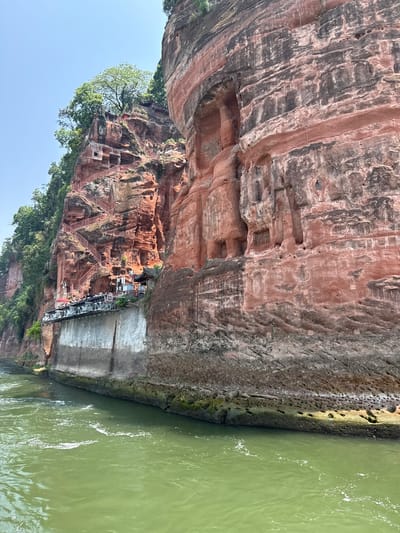 The image depicts a section of the Leshan Giant Buddha scenic area in Shizhong District, Leshan, Sichuan, China, specifically the cliff face adjacent to the river. The cliff is primarily composed of red sandstone with visible weathering and erosion patterns. The image shows a complex of carved structures including a series of stairways constructed along the cliff face, leading to elevated platforms and structures. The steps are painted red and appear to be made of metal. The environment is lush, with trees growing on the cliff. Green river water is visible at the base of the cliff.