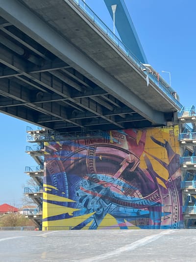 The image displays a large concrete and steel bridge structure in Medgidia, Romania, viewed from beneath. A substantial concrete bridge pier in the center is covered by an extensive, multi-colored mural. The mural features abstract, geometric designs resembling interconnected gears, compasses, and numerical scales, utilizing vibrant blues, purples, yellows, and reds. Text reading "SWEET DAMAGE RECKS" is visible on the upper-right section of the mural. Multiple external metal staircases with blue railings are affixed to the sides of the pier. The underside of the bridge deck reveals concrete slabs, metal girders, and pipes. The upper portion of the bridge features blue guardrails and a prominent blue diagonal suspension component against a clear blue sky. The ground beneath the bridge is an asphalt or concrete surface with a partial white line. To the distant left, sparse trees and a red-roofed building are visible. No individuals are present in the scene.