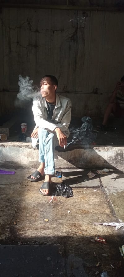 An individual, identified as a young adult male, sits on a raised concrete ledge in what appears to be a dimly lit, dilapidated, and cluttered semi-enclosed area within Sulaeman, Indonesia. He is positioned facing slightly right, exhaling a significant cloud of smoke from a cigarette held between his lips. His left hand rests on his lap, while his right hand holds another cigarette. He wears a light-colored jacket, a dark t-shirt, light blue jeans, and black slide sandals. Strong sunlight illuminates the front of his face and parts of the floor in front of him. To his left, on the same ledge, is a white disposable cup with red markings and some debris. To his right, on the ledge and ground, there are several discarded items, including crumpled black plastic, cigarette butts, and paper. Another person is partially visible in the deep background to the right, sitting on the ground in striped clothing. The background features a textured, cracked, and stained concrete wall. The concrete floor in the foreground is dirty, wet in areas, and strewn with various trash items, including more cigarette butts and torn paper.