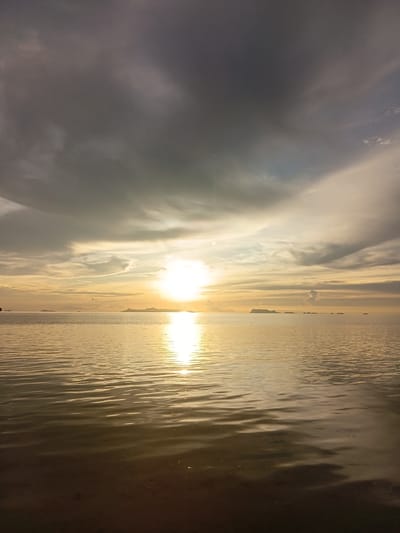 The image depicts a serene seascape at sunset, likely in Baan Pang Ka, Thailand. The sun, a brilliant orb, is low on the horizon, casting a warm, golden glow across the tranquil water. Wispy clouds, tinged with orange and yellow, fill the sky, creating a dramatic contrast with the darker, heavier clouds that loom overhead. The water's surface is gently rippled, reflecting the colors of the sky and the sun's intense light. In the distance, several islands are silhouetted against the bright horizon, their forms softened by the atmospheric haze. The foreground shows the shallow, dark water, with hints of the sandy bottom visible. The overall impression is one of peaceful solitude and natural beauty.