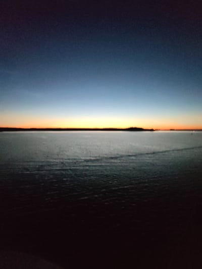 The image depicts a body of water, likely a lake or sea, at dusk. The sky transitions from a deep indigo at the top to lighter blues and then a thin band of orange and yellow near the horizon, indicating the setting sun. A dark, silhouetted landmass, possibly a shoreline with trees, forms a line across the horizon. The water surface is dark and appears somewhat choppy, with subtle ripples visible. In the foreground, the water darkens significantly, eventually becoming almost black. There are no people or discernible objects in the immediate scene. The overall impression is one of a serene, natural landscape at the end of the day. There is no indication of the city of Irdomatka, Russia, or any urban elements within the image.