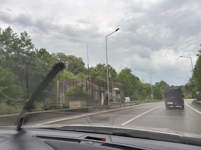 An overcast, grey sky looms above a wet, asphalt road in Buknari, Georgia. Trees line the left side of the road, and a concrete structure with a fence sits next to it. A black van drives on the right side of the road. The image is taken from inside a car, with the windshield wiper visible in the foreground.