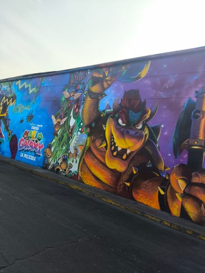 The image shows a mural on a wall adjacent to a street in the La Victoria district of Lima, Peru. The mural depicts characters and imagery from the "Super Mario Galaxy" video game series, with a focus on Bowser. The mural includes the text "Super Mario Galaxy: La Pelicula," suggesting a movie adaptation concept. The background behind Bowser fades into a purple starry sky. The mural appears to be painted on a rough textured wall next to the road.