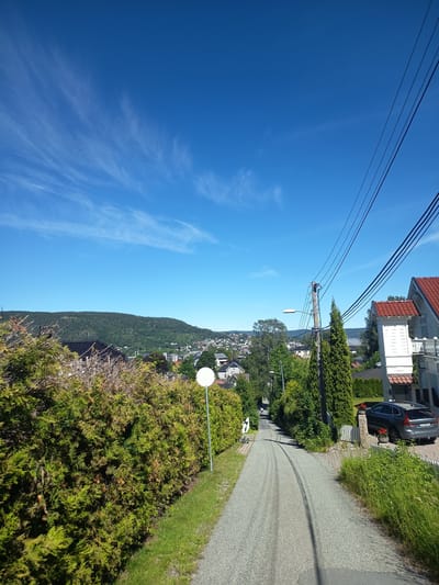 A high-angle, wide shot shows a residential street winding uphill in Drammen, Norway. The sky is bright blue with wispy white clouds. Lush green trees and hedges line the street, with houses visible in the background. A grey SUV is parked in a driveway on the right side of the road. The street surface is gravel with tire tracks. Electrical wires run overhead, supported by a utility pole.