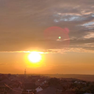 This image captures a sunset over Tohatin, Moldova. The sky is filled with warm hues of orange and yellow, with scattered clouds. The sun, a brilliant orb, is partially obscured by a thin layer of clouds on the left side of the frame, casting a bright glow. In the upper right quadrant of the sky, a fainter, green-tinted circular lens flare is visible.
Below the sky, a silhouette of a town or village is visible. Rooftops of houses, interspersed with trees, form a dense pattern. A prominent lattice-like transmission tower stands tall on the left side of the horizon line. The landscape appears somewhat hilly, with a gentle slope leading into the distance. Two small birds are visible in flight against the expansive sky. The overall impression is one of a tranquil evening in a semi-urban or rural setting.