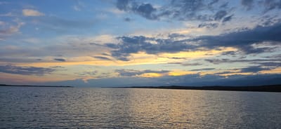 The image shows a wide expanse of water, likely a lake or sea, under a dramatic sunset sky. The water is calm with gentle ripples, reflecting the colors of the sky. The sky is a canvas of blues, grays, and yellows, with clouds forming interesting patterns as the sun sets. A distant shoreline is visible, with a low, dark silhouette of land and mountains against the horizon. The overall atmosphere is serene and picturesque, characteristic of a sunset over a large body of water. Given the location provided, this scene likely depicts Issyk-Kul Lake near the city of Pristan'-Przheval'sk (Karakol), Kyrgyzstan, a popular destination known for its stunning natural beauty and sunsets. There are no people or specific objects clearly identifiable in the image, focusing solely on the natural landscape.