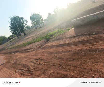 This image depicts a dirt road in what appears to be a rural area, potentially in or near Wase, Nigeria, given the context. The foreground is dominated by a reddish-brown dirt surface marked with tire tracks. Patches of sparse vegetation are visible along the edges of the road, and a concrete barrier or wall sits along the side of the road on the right. The background includes trees scattered across a gentle slope under a bright sky with sunlight. There are no people visible in the image.