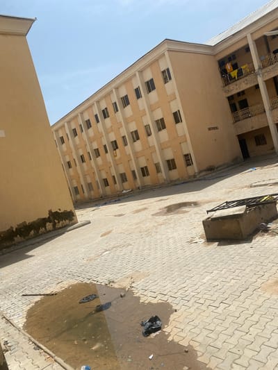 This image shows an outdoor courtyard area with two large buildings. The building on the left is a light tan color with peeling paint near the bottom. The building on the right is a peach color with white trim and a metal roof. It has multiple stories with balconies and numerous windows. Some clothing, appearing to be yellow and white, is hung out to dry on a balcony on the right side of this building. A sign on the peach-colored building reads "CHRISTOPHER DEGA". The ground is paved with light-colored interlocking bricks. Several puddles of muddy water are present on the paving, with some debris, including black plastic bags, scattered around. In the foreground to the right, there is a concrete structure with a metal frame on top, and more debris around it. The sky is clear and blue. The scene appears to be in an urban or semi-urban environment. This location is identified as being in the city of Yan Dala, Nigeria.