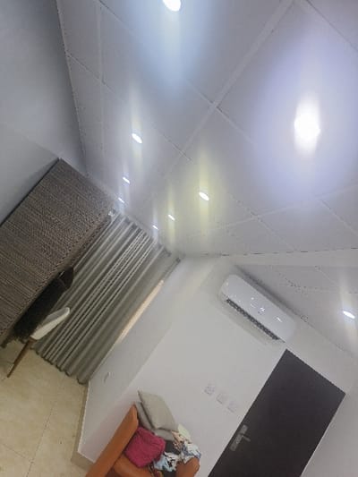 The image depicts an interior room in Close, Nigeria, viewed from an elevated, tilted perspective. The room features a white, square-tiled ceiling equipped with multiple recessed, illuminated spotlights. Walls are uniformly white. In the upper left, a dark brown wooden cabinet with horizontal grain is positioned against a wall, partially obscuring a light-colored chair. Behind these objects, a window is covered by vertical grey blinds. On the opposite wall to the right, a white split-unit air conditioner displaying "26" is mounted high. Below it, three white electrical outlets are horizontally aligned. A dark-colored door with a silver handle is visible on the far right. In the lower right, an orange or tan upholstered sofa or armchair contains a light grey cushion, a pink fabric item, and a patterned fabric item. The floor in the lower left consists of light-colored tiles. No individuals are present in the scene.