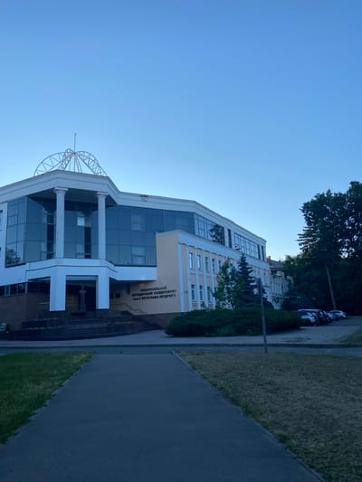 The image depicts the National Law University named after Yaroslav the Wise in Nyzhni Mlyny, Ukraine. It is a bright, clear day, likely late afternoon or early evening, given the angle of the sunlight. The main subject is the university building, a modern structure with large glass windows and white architectural elements, including columns and a prominent facade. The building is surrounded by greenery, including manicured hedges and trees. A paved walkway runs alongside the building, and a strip of grass is visible. In the background, several cars are parked along a street lined with trees. The sky is a clear, vibrant blue. The text on the building reads "НАЦІОНАЛЬНИЙ ЮРИДИЧНИЙ УНІВЕРСИТЕТ ІМЕНІ ЯРОСЛАВА МУДРОГО" which translates to "NATIONAL LAW UNIVERSITY NAMED AFTER YAROSLAV THE WISE". There are no people visible in the image. The overall impression is one of a serene and well-maintained educational institution.