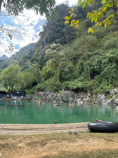 A vibrant landscape with a serene lake, surrounded by lush green mountains and foliage. A bamboo raft extends from the grassy shoreline into the emerald water, holding a black inner tube. Several people are swimming in the lake near the rocky edge. A building sits near the water's edge, nestled among the trees. The scene captures a peaceful, natural environment.