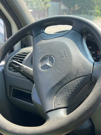 This is a close-up, eye-level, medium shot of a Mercedes-Benz steering wheel in a vehicle. The steering wheel is dark gray, made of textured plastic, and features the Mercedes-Benz logo in the center. The "SRS AIRBAG" text is visible on the right side of the airbag cover. To the left of the steering wheel, a black turn signal lever with light symbols is partially visible, along with air vents. The dashboard is also gray. In the background, through the windshield, blurred greenery and a building can be seen, suggesting an outdoor, possibly sunny environment. There are no people present in the image. The image was taken in Khust, Ukraine.