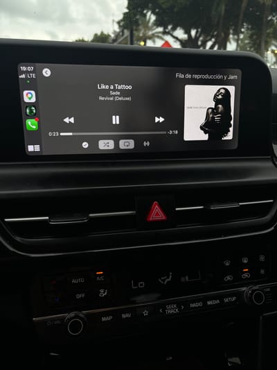 Here is a description of the image:
The image shows the interior of a car, focusing on the infotainment system and center console. The infotainment system displays a music player interface with the song "Like a Tattoo" by Sade from the "Revival (Deluxe)" album. The song is 0:23 seconds in and has 3:18 remaining. The display is connected to an iPhone via Apple CarPlay and is in a "Fila de reproducción y Jam" playlist, featuring Sade Love Deluxe album art. The center console has climate control buttons (A/C, Auto, Off), a hazard light button, and audio controls (Map, Nav, Seek Track, Radio, Media, Setup, Tune/Volume). The car is likely in a parking lot or stopped on the street as the background shows trees and street lights. No people are visible in the image. Based on the car model's dashboard design and the environment in the background, the image appears to be taken in a Kia vehicle. While the image doesn't explicitly show Lauderdale-by-the-Sea, United States of America, the presence of "LTE" connection signal suggests that the car is most likely in a modern city in United States where LTE internet service is provided.