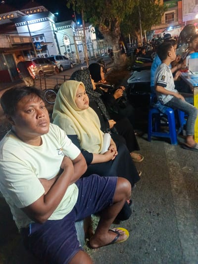 The image depicts a street scene at night, likely in Luwoo, Indonesia, with several people gathered. In the foreground, a man wearing a light yellow t-shirt and dark blue patterned shorts sits with his arms crossed, looking off to the right. Beside him, a woman in a yellow hijab and a dark patterned garment is also seated. Further back, more individuals are visible, some seated at tables, suggesting a food stall or dining area. A mosque with white architecture and arched windows is visible in the background, illuminated by lights. Several vehicles, including cars and motorcycles, are parked along the street. The overall environment appears to be a bustling urban night setting with artificial lighting casting shadows.