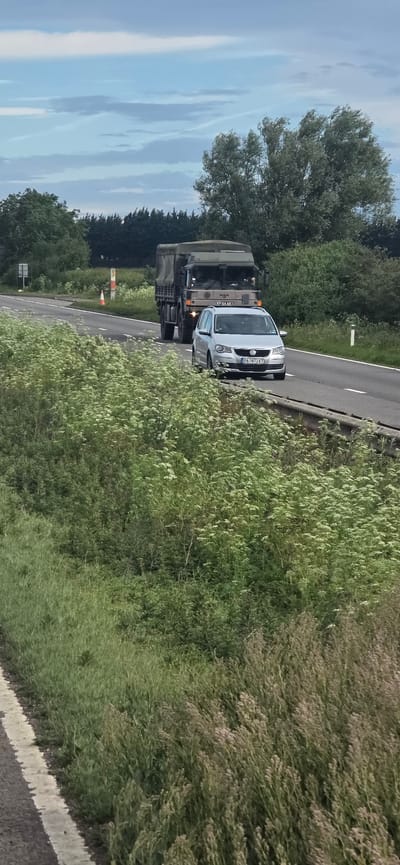 A silver Volkswagen Touran is driving on a rural road in South Kesteven, United Kingdom, passing a military truck. The Volkswagen has license plate PA7873KT. The military truck appears to be a MAN truck, with license plate KP64 AB. The road is flanked by lush green foliage and trees. The sky is overcast with scattered clouds. The scene suggests a typical day on a rural highway in the UK.