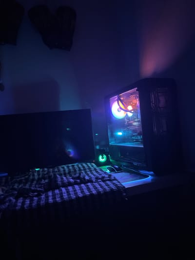 The image depicts a dark room illuminated by the internal RGB lighting of a desktop computer case. The computer case is situated atop a black cabinet or tower. To the left of the cabinet, there is wall decor with what appears to be three black patches, likely wall art. To the right of the cabinet, hanging vertically, is a patterned cloth or garment, possibly plaid. The room has a blue and purple hue due to the computer's lighting. There are no people visible. The location, Bayraklı, Turkey, cannot be confirmed from the provided visual information.