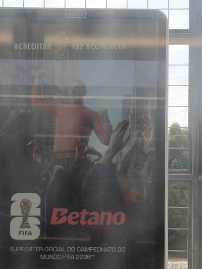 A man in an orange jacket is shown raising his fist in a celebratory gesture, surrounded by other people's hands reaching towards him. The man appears to be on a stage or elevated platform, as suggested by the background which includes a cityscape and the FIFA World Cup trophy. Text on the poster reads "ACREDITAR FAZ ACONTECER" and "Betano," along with "SUPORTER OFICIAL DO CAMPEONATO DO MUNDO FIFA 2026". The poster is displayed on a public transport stop in Palmela, Portugal, as indicated by the "MOP" logo, which stands for Mobilidade Ocupação Participação, a municipal program in Palmela. The poster is framed by a window with metal grating, and outside the window, trees and power lines are visible, suggesting an urban or suburban setting. The image is captured during daylight, with sunlight casting reflections on the poster's surface.