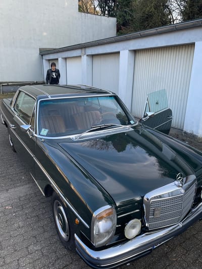The scene takes place outdoors in Oberhausen, Germany. A dark green Mercedes-Benz W114/W115 coupé is prominently featured, parked on a dark grey paver surface. The vehicle is angled towards the viewer's right, showcasing its front right quarter. It possesses a reflective, polished finish, chrome bumpers, a chrome grille with the Mercedes-Benz star emblem, rectangular headlights, and circular driving lights. The interior, visible through the windshield and the open passenger-side door, displays brown pleated leather upholstery and a wooden-rimmed steering wheel. A male individual with dark hair, clad in a black leather jacket and a black t-shirt with a white graphic, stands behind the car, facing forward. He is positioned in front of a series of at least three white vertically-ribbed garage doors, with numbers "15" and "16" partially visible. Above the garage doors, a light grey building wall and leafless tree branches are visible against a bright sky.