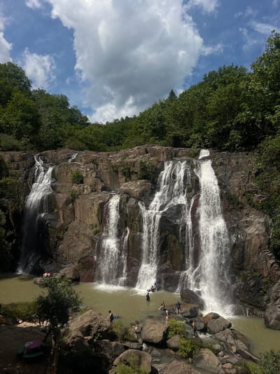 The image depicts a multi-tiered waterfall cascading down a rocky cliff face, surrounded by lush green trees under a cloudy blue sky. Several people are visible in and around the water at the base of the falls, with some swimming or wading, and others relaxing on the rocks. One person is lying on a large rock near the left side of the pool, and another person is standing in the water on the right side, partially obscured by a rock. There is a picnic table with benches visible in the lower left corner of the frame. The scene appears to be a natural recreational area, potentially a park or a designated swimming spot. The specific location in Arambala, El Salvador is not explicitly indicated by any visual cues within the image itself.