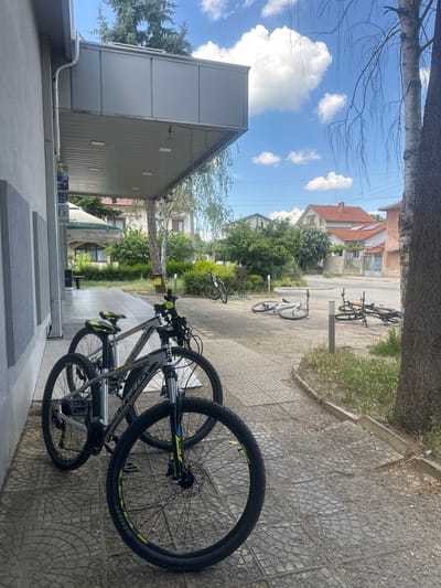 In Ovoshtnik, Bulgaria, two bicycles are parked on a paved walkway in front of a building with a gray, modern overhang. The bicycles are positioned side-by-side, with one slightly in front of the other. The foreground bicycle is a mountain bike with a gray frame, black handlebars with yellow grips, and black tires with yellow accents. Its front wheel is turned slightly to the left. Behind it, another similar bicycle is partially visible. The walkway is made of gray interlocking pavers, with some scattered debris and dry grass along the edge. To the right, a large tree trunk with rough bark stands tall. In the background, a residential street is visible with houses featuring red-tiled roofs and green trees. The sky is blue with scattered white clouds. Further down the street, additional bicycles are seen lying on the ground.