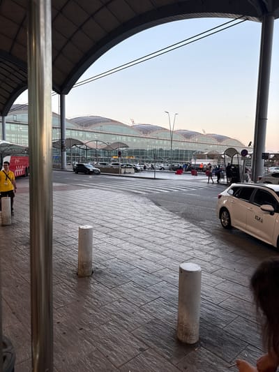 The image depicts the exterior of Alicante–Elche Airport in Elx (Elche), Spain. The foreground features a tiled pavement with several cylindrical bollards. A person wearing a yellow shirt and glasses stands near a red bus on the left side of the frame. Several vehicles, including cars and what appears to be a taxi with "ELX" branding on its side, are visible in the middle ground and background. The airport terminal building, characterized by its glass facade and undulating roof structure, dominates the background. The scene is illuminated by natural daylight, suggesting it was taken during the day.