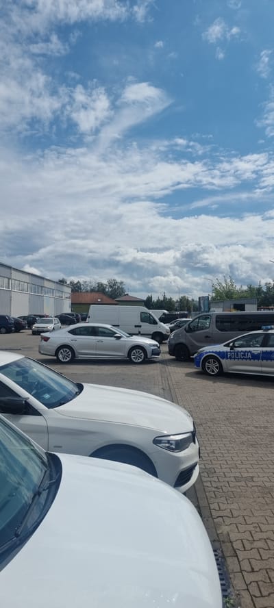 In Mroków, Poland, a parking lot adjacent to a commercial building is the setting. A fleet of vehicles is parked on a paver surface under a partly cloudy sky. In the foreground, the hood and windshield of a white vehicle are visible. Behind it, a silver sedan is parked. To the right of the sedan, a police car with "POLICJA" text on its side is positioned. Other vehicles, including vans and sedans, are visible throughout the parking lot. A low-rise building with a metallic facade is located to the left, with a tiled-roof building visible behind it. Trees and utility poles are interspersed in the background.
