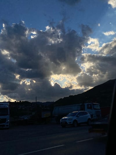 The image depicts a cloudy sky with sunlight breaking through the clouds in Prijepolje Municipality, Serbia. In the foreground, a paved road is visible with a white SUV parked on it. To the left, a white truck is parked, and behind it, a residential area with houses built on a hillside can be seen. To the right, a dark mountain silhouette is present. The overall scene suggests a late afternoon or early evening setting due to the dramatic lighting from the sun. There are no people visible in the frame.