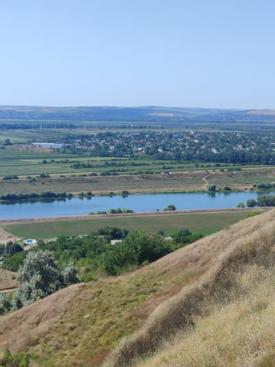 An aerial, eye-level, full shot captures a serene landscape on a clear day in Bălăbănești, Moldova. The sky is a bright, cloudless blue. Below, a vast expanse of rolling hills covered in dry, golden grass slopes down towards the foreground. A winding dirt path is visible on the side of the hill.
In the middle ground, a wide, calm river with a blue surface reflects the sky. Along the riverbanks, there are patches of green grass and trees. Beyond the river, a town or village is nestled amongst lush green trees, with houses featuring red and white roofs. Agricultural fields, mostly green, are interspersed with more trees, creating a patchwork effect.
Further in the background, more distant hills are visible, some with a yellowish hue, suggesting drier terrain. The overall impression is one of a peaceful rural environment, showcasing the natural beauty and settlement patterns of the region. The image is composed with a slight tilt, giving it a dynamic feel.