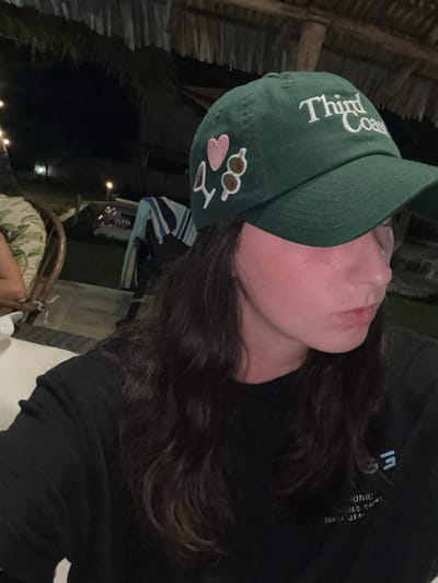 A woman with long, dark, wavy hair wears a dark green baseball cap with the words "Third Coast" embroidered on it, plus patches of sunglasses, a heart, and an "I heart NY" design. She wears a black long-sleeved shirt and is seated in a wicker chair. The background shows a dimly lit outdoor setting with a thatched roof and patio furniture. The overall lighting is low, creating a slightly grainy effect.