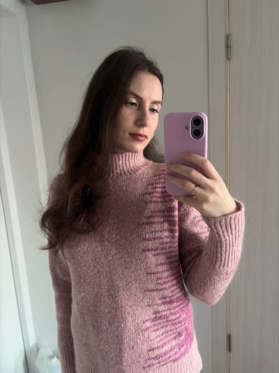 A woman in a pink and purple sweater takes a selfie in a mirror. She holds a lavender phone, the camera side facing forward. She has long brown hair and is wearing a ring on her ring finger. A small portion of a white wall and door are visible behind her.