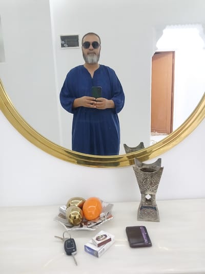 This media file is a mirror selfie taken in a room. The subject is a man with a beard and sunglasses, wearing a blue robe. He is holding a smartphone and taking the picture. In the reflection, we can see a window with a wooden frame at the top of the room, and a small framed artwork on the wall. On a shelf to the right of the mirror, there are several objects, including a decorative metal incense burner, a dark brown wallet with a purple card peeking out, a small dish with three decorative spheres (one orange and two gold), a pack of cigarettes, and a car key. The setting appears to be indoors, likely a home. The lighting suggests it is daytime. The overall mood is casual and personal.