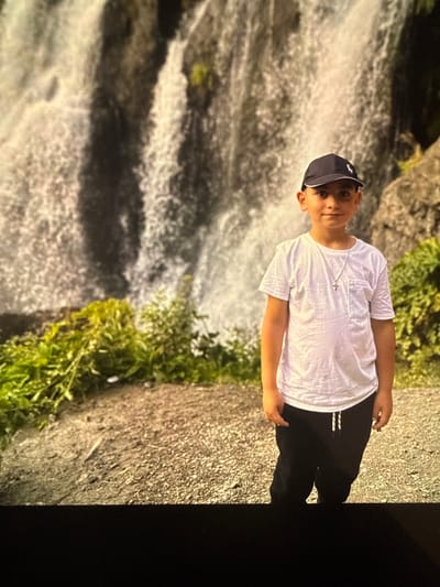 A young boy wearing a white t-shirt, black pants, and a dark baseball cap stands in front of a waterfall. The boy is the central subject of the image, positioned in the foreground. He is looking towards the camera with a neutral expression. He is wearing a silver necklace with a cross pendant. The waterfall dominates the background, appearing wide and textured with water cascading down. Green foliage is visible to the left and right of the boy, near the base of the waterfall. The ground in front of the boy appears to be gravelly. The scene suggests an outdoor natural environment, possibly a tourist attraction in or near Artik, Armenia, given the presence of waterfalls in the region.