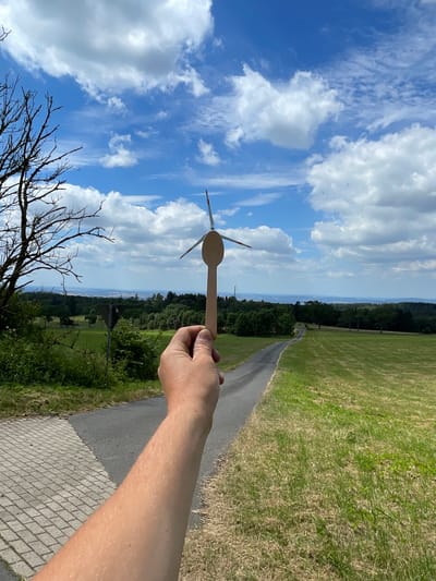 A person's arm and hand are in the foreground, holding a wooden spoon with a miniature wind turbine attached to its bowl. The wind turbine has three blades, two of which are visible and tipped with red. The scene is outdoors, with a paved path on the left and a grassy field on the right, leading towards a winding asphalt road in the middle ground. Trees and rolling hills form the background, under a partly cloudy blue sky. The location is identified as Schwarzenborn, Germany.