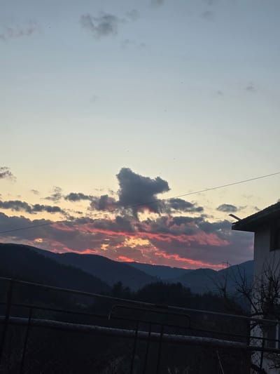 A landscape image, taken from what appears to be a balcony or elevated position, oriented vertically. The sky features a vibrant sunset with pink and orange hues interspersed with gray clouds. A dark cloud formation is centered within the sky. The background reveals a mountain range. To the upper right, portions of a building structure are visible. In the foreground, a metal railing is seen. The location cannot be confirmed to be Giovren, Bulgaria as no information is provided to that effect in the image.