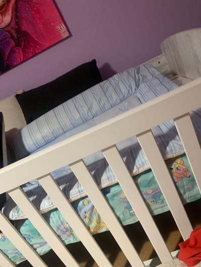 An indoor, eye-level shot depicts a crib with a purple wall and a large painting of a character in pink and purple on the left. The crib is adorned with light blue and white striped bedding. A long, dark gray pillow rests at the head of the crib. Beneath the striped bedding, a teal sheet with cartoon characters, including one resembling a pink pony and another a blue creature, is visible. The crib is made of white wood, with vertical slats. A small portion of a red garment is visible in the lower right corner. The image was taken inside a home in Krushev Dol, Bulgaria.