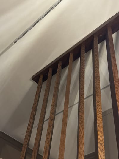 A low-angle shot presents modern wooden stairs affixed to a white wall. The stairs consist of horizontal, parallel wooden planks, with dark grain visible. The structure is attached diagonally to the wall. A silver metal strip accents the wall's left side. The lighting is soft, creating subtle shadows on the wall and stairs.