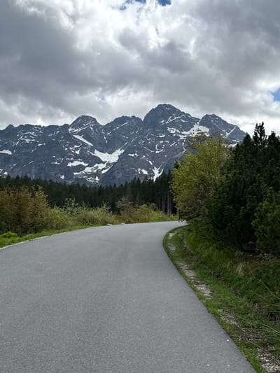 The image shows a road in a mountainous area. The road is asphalt and appears to be well-maintained. It curves slightly to the right. On the right side of the road is a grassy verge with some vegetation. Beyond the road, a tall, snow-capped mountain dominates the landscape. The mountain is dark rock with patches of snow clinging to its sides. The sky is cloudy, with grey clouds obscuring the peaks. The location is not in the city of Brzegi, Poland, based on the mountainous landscape and absence of urban features.