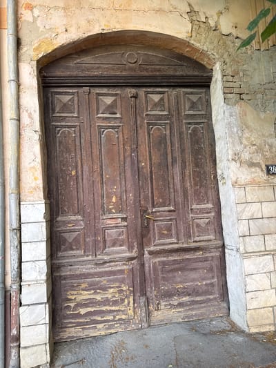 The image depicts an aged, double-leaf wooden door set within a weathered arched doorway, located in Becej Municipality, Serbia. The door, painted a dark maroon or brown, exhibits significant peeling and flaking paint, revealing lighter wood beneath, particularly at the bottom panels. Each door leaf features four distinct panels: two elongated vertical panels with arched tops and two square panels, with the upper square panels showcasing raised pyramidal centers. A brass-colored doorknob and keyhole are present on the left door leaf.

The arched doorway surround is constructed from materials like brick or stone, heavily coated with plaster that is extensively cracked, flaking, and deteriorated, exposing layers of yellowed undercoat, bare brick, and possibly original stone. Above the door, within the arch, there is a subtle carved decorative element, including a central circular motif.

To the immediate left of the doorway, a vertical silver-colored metal drainpipe runs down a wall of light, cracked, and peeling plaster, which transitions into a lower section of neatly stacked rectangular light-colored tiles. On the right side of the doorway, the wall above is severely damaged, exposing red brickwork and rough plaster, while the lower section mirrors the left side with similar light-colored rectangular tiles. A black rectangular house number plate displaying "38" in white digits is affixed to the tiled section on the right. A few green leaves are barely visible at the top right, suggesting nearby foliage. The ground directly in front of the door is a dark, rough-textured surface, possibly concrete or asphalt, with some minor debris. No people are present in the scene.
