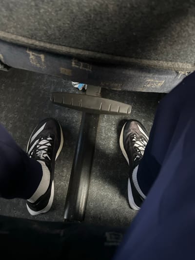 The image depicts an overhead view of a person's lower legs and feet within an enclosed public or commercial setting located in Richmond, United States. The individual is wearing dark blue trousers and white socks. Both feet are shod in black and white Adidas Adizero athletic shoes, featuring white outsoles and prominent white stripes on the lateral sides, with "ADIZERO" text visible on the tongue of the left shoe. The feet rest on a dark, speckled floor surface. Centered between the subject's feet is a black, vertical cylindrical pole with a horizontal T-shaped footrest attached at its base, positioned on the floor. Directly behind the footrest, beneath a darker blue upholstered surface (likely a seat or barrier), a crumpled piece of white and blue litter is visible. The upper portion of the image reveals a dark grey, textured surface, possibly the underside of a seat or a wall panel, with visible wear and lighter markings. The overall environment suggests public transportation or a stadium/arena seating area.