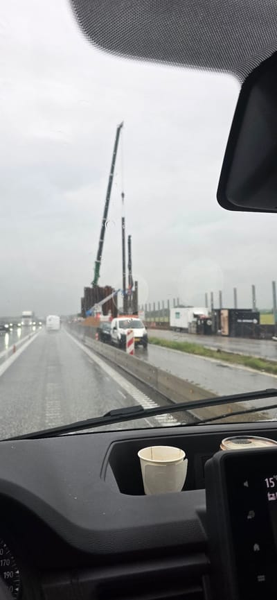 The image, taken from inside a vehicle, shows a construction site on a rainy day on what appears to be a highway near Hatting, Denmark. A large crane is visible, lifting a tall, dark pole. Several other vertical structures are in the background, suggesting the ongoing construction of a bridge or similar infrastructure. The road is wet and shows lane markings, with other vehicles in the distance. In the foreground, inside the vehicle, a disposable paper cup sits in a cup holder on the dashboard. A digital display screen is also visible to the right. The overall impression is of a functional, work-in-progress environment despite the inclement weather.