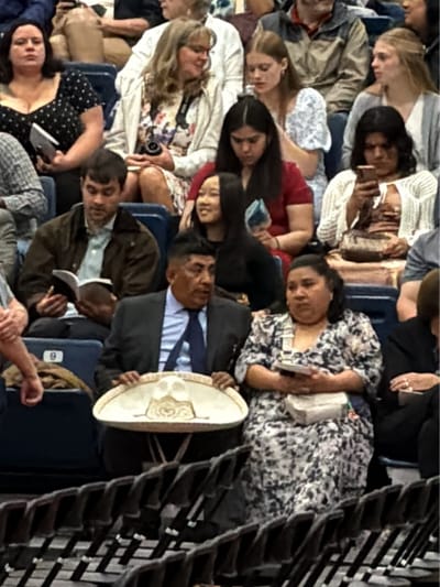 The image shows a group of people seated in an auditorium-style seating arrangement. A man in a suit and tie holds a large, decorative sombrero on his lap. Seated next to him is a woman in a floral patterned dress with a small purse on her lap. Other individuals are seated in rows around them. One man is reading a book. People can be seen using phones and cameras. The specific location within the city of Rochester is not discernible from the visual details provided.