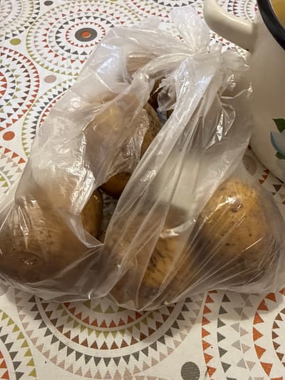The image shows a plastic bag filled with potatoes resting on a patterned tablecloth. The tablecloth has a repeating geometric design featuring triangles and circles in shades of white, cream, orange, brown, green, and gray. Part of a white ceramic bowl or pitcher with a black rim is visible in the upper left corner of the image, suggesting it might be used to hold or serve the potatoes. The potatoes themselves are light brown and appear to be raw, with some dirt or imperfections visible on their skins. The lighting suggests it is daytime, and there are no specific cues to indicate the weather or a particular emotion. The scene appears to be indoors, likely in a kitchen or dining area. There is no visible text in the image. The location context of Morozovka, Russia is provided, but there are no visual elements within the image that specifically confirm this location.