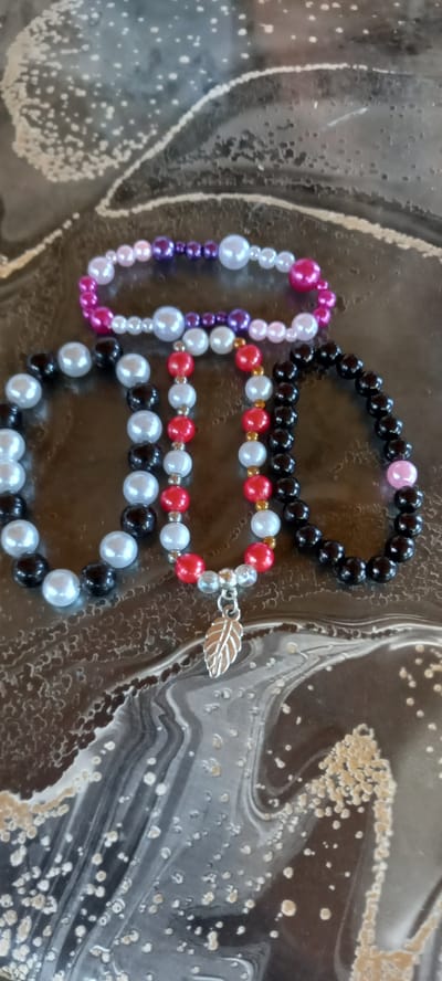 This is a close-up, top-down shot of four handmade beaded bracelets arranged on a dark, marbled surface. The bracelets are laid out in a fanned formation, with the top bracelet a gradient of pink and purple pearls. Below it, a bracelet of white and purple pearls, and then a bracelet of black and white pearls. The lowest bracelet is red and white with a gold accent bead and a small, silver leaf charm dangling from it. The surface beneath appears to be a glossy, dark countertop with abstract, swirling patterns in white and beige. The image is taken from a high angle, focusing on the bracelets. There is no indication of the city of Iskra, Bulgaria, in the image.