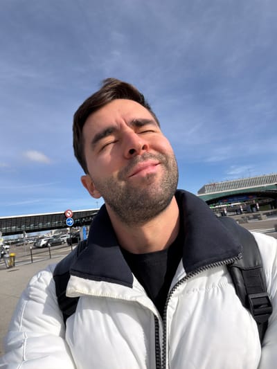 A male individual is depicted in a selfie, taken outdoors on a sunny day at Lyon-Saint Exupéry Airport in Colombier-Saugnieu, France. The man, with short dark hair and a beard, is shown with his eyes closed and head tilted slightly upward, exhibiting a serene expression as if exposed to sunlight. He is wearing a white puffer jacket with a black collar over a black t-shirt, and a black backpack strap is visible over his right shoulder. The background features airport infrastructure: a paved roadway or drop-off area with numerous parked vehicles, an elevated walkway structure on the left, and a large terminal building complex with multiple overhead gantries and partially visible green signage on the right. Two circular road signs, one red with a white horizontal bar and another blue with a white right-pointing arrow, are mounted on a pole in the mid-ground to the left. The sky above is clear blue with minimal, wispy white clouds.
