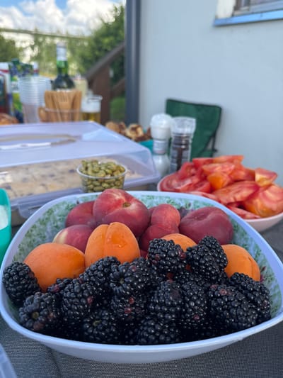 An outdoor dining scene is captured on a table with a grey tablecloth. In the foreground, a white bowl with a green floral pattern holds a generous serving of fresh blackberries and several ripe apricots. Behind the fruit bowl, a clear plastic container with a lid displays what appears to be a baked good, possibly a cake or pastry, covered in a dusting of powdered sugar. To the right of the cake, a small clear plastic cup is filled with green pistachios. Further back on the table, a white bowl is filled with sliced red tomatoes. A salt and pepper grinder set is visible near the tomatoes. In the background, a green camping chair is partially visible. A blurred bottle of beer and other indistinct items are on the table and a wooden railing is present. The sky above is partly cloudy. The overall impression is a casual outdoor meal.