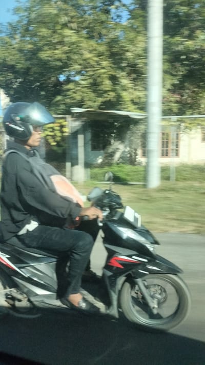 A person is riding a black motorcycle on a paved road. The rider is wearing a black helmet, sunglasses, a dark grey long-sleeved shirt, and dark pants. They are also wearing a bright orange vest over their shirt. The motorcycle has red and white decals. In the background, there are trees, a metal pole, and buildings with windows. The scene appears to be in Blang Seupeung, Indonesia, given the general appearance of the vegetation and buildings, and the presence of a motorcycle, a common mode of transportation in the region. The image is blurred due to motion, suggesting it was taken from another moving vehicle.