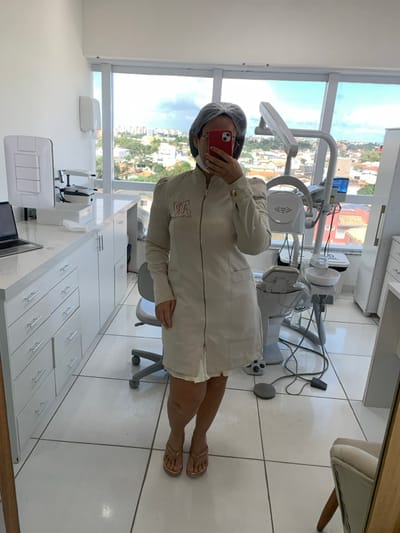 The image depicts a well-lit, modern dental or medical treatment room located in the city of Lauro de Freitas, Brazil. A single individual, presumed female, stands centrally, taking a mirror selfie with a red iPhone that obscures her face. She is dressed in a light-colored, long-sleeved medical coat or tunic featuring a front zipper and an embroidered "SPA" logo (or similar stylized text) on the left chest. Her head is covered by a dark hairnet, and she wears gold-colored thong sandals.
The room has white tiled flooring with grey grout lines and white walls. A large window covering the back wall provides a clear view of an urban environment in Lauro de Freitas, characterized by a mix of buildings, trees, and a partly cloudy daytime sky. On the left, a white counter with multiple drawers supports a silver laptop, a medical imaging device, and a disposable face mask. A wall-mounted dispenser is also present. On the right side, a white dental chair with an integrated unit, including a cuspidor bowl, light, and various dental instruments, is prominent. A grey, wheeled medical stool and a foot pedal are on the floor near the dental unit. A portion of a wooden-framed mirror, reflecting a beige upholstered chair, is visible on the far right.