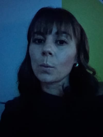 A woman with shoulder-length dark hair and bangs looks at the camera. She is fair-skinned and wearing a dark shirt. There appears to be a dark earring. The background is divided into three distinct sections: a blue area at the bottom left, a green area at the top left, and a dark area at the right. The lighting is dim, with a bluish tint overall.